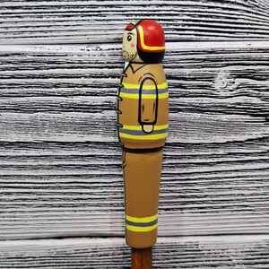 May include: A wooden figurine of a firefighter wearing a red helmet and a brown and yellow uniform. The figurine is painted with details, including a face, a mustache, and a black belt.