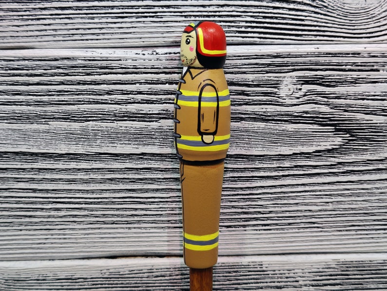 May include: A wooden figurine of a firefighter wearing a red helmet and a brown and yellow uniform. The figurine is painted with details, including a face, a mustache, and a black belt.