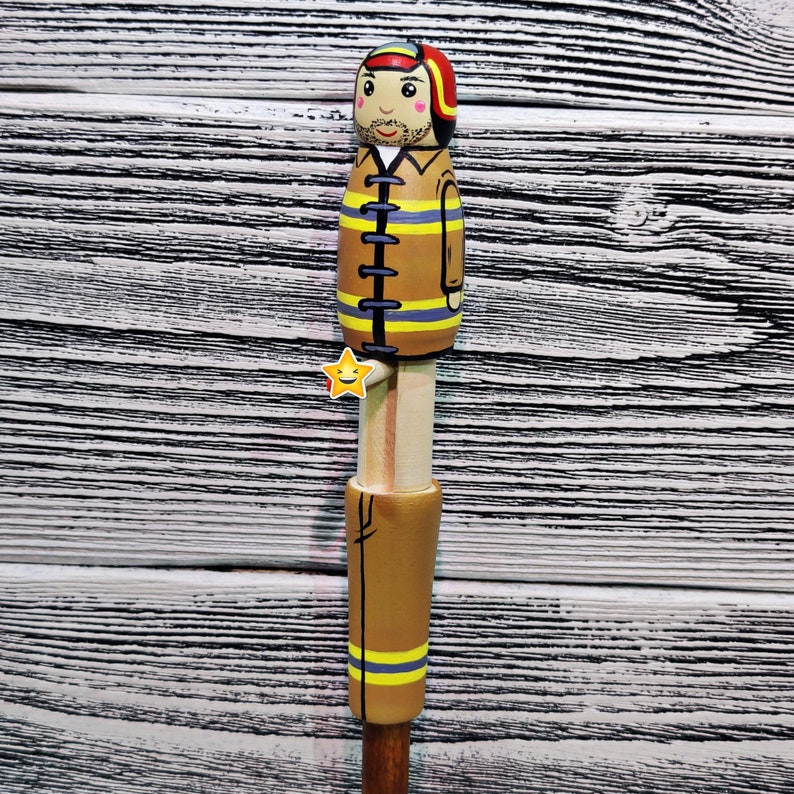 May include: A wooden figurine of a firefighter wearing a yellow and brown uniform with a red helmet and a smiling face. The figurine is attached to a wooden stick.