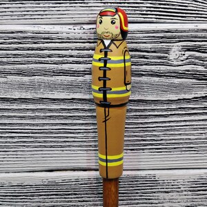 May include: A wooden figurine of a firefighter wearing a brown uniform with yellow stripes, a red helmet, and a black belt. The figurine is painted with a realistic face and has a mustache.