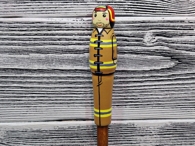 May include: A wooden figurine of a firefighter wearing a brown uniform with yellow stripes, a red helmet, and a black belt. The figurine is painted with a realistic face and has a mustache.