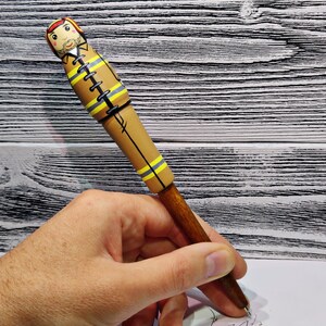 May include: A wooden pen with a brown and yellow painted design of a firefighter. The pen is held in a hand.