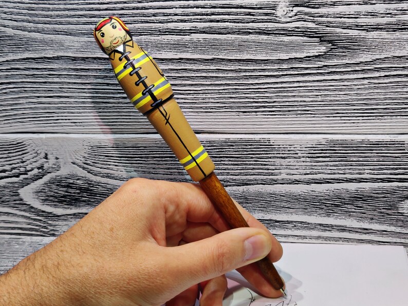 May include: A wooden pen with a brown and yellow painted design of a firefighter. The pen is held in a hand.