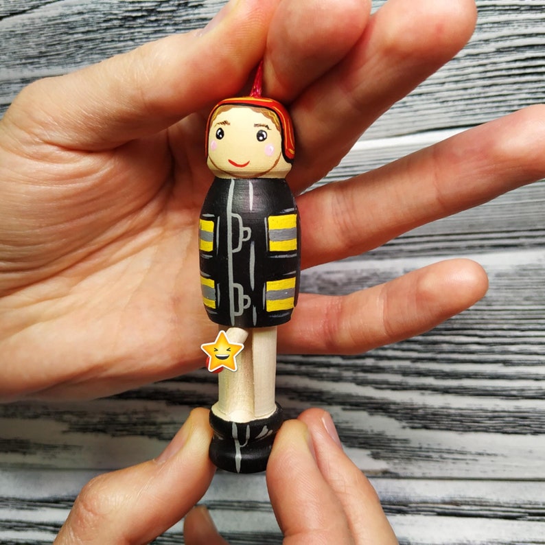 Pode incluir: Figurinha de brinquedo de madeira de um bombeiro vestindo um uniforme preto e amarelo com um capacete vermelho. A figurinha tem um rosto pintado com uma express&atilde;o sorridente e um adesivo de estrela amarela no peito.