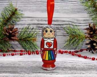 May include: A wooden Christmas ornament shaped like a person with gray hair, wearing a red dress with a rainbow skirt and a red heart on the chest. The ornament has a red ribbon for hanging.