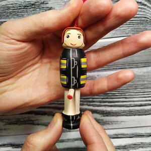 Pode incluir: Uma figurinha de madeira pintada &agrave; m&atilde;o de um bombeiro vestindo um uniforme preto e amarelo com um capacete vermelho. A figurinha tem um rosto sorridente e est&aacute; segurando um pequeno objeto vermelho.