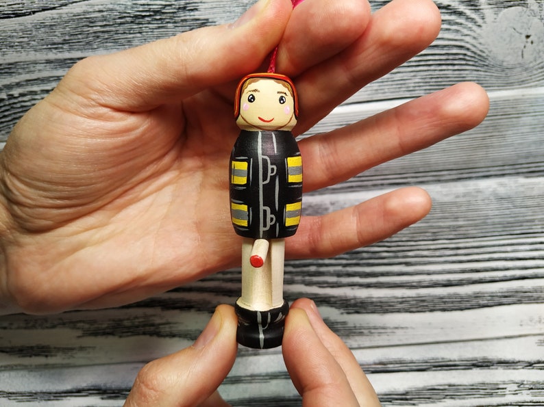 Pode incluir: Uma figurinha de madeira pintada &agrave; m&atilde;o de um bombeiro vestindo um uniforme preto e amarelo com um capacete vermelho. A figurinha tem um rosto sorridente e est&aacute; segurando um pequeno objeto vermelho.