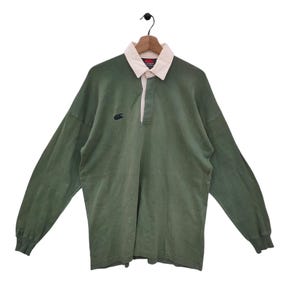 Puede incluir: Una camiseta de rugby de manga larga verde con cuello blanco y un pequeño logotipo negro en el pecho izquierdo. La camiseta tiene una abertura de botones y un cuello de canalé.