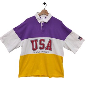 May include: A short-sleeved rugby shirt with a color-block design. The top is purple, white, and yellow, with "USA" in red block letters across the chest. The text "THE STARS AND STRIPES" is below the USA text. An American flag patch is on the sleeve.
