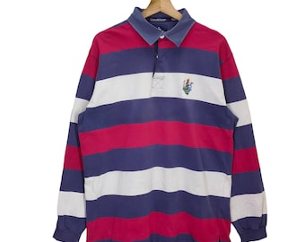 May include: A long-sleeved rugby shirt with a striped pattern in navy blue, white, and red. The shirt has a button-up collar and a small embroidered logo on the chest.