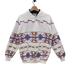 Könnte beinhalten: Weißer Langarm-Pullover mit geometrischem Muster in Violett-, Rot- und Gelbtönen. Der Pullover hat einen Knopfkragens und eine Tasche auf der linken Seite.
