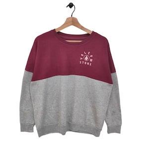 May include: A maroon and grey color block sweatshirt with long sleeves and a crew neck. The sweatshirt has a Volcom Stone logo on the front.