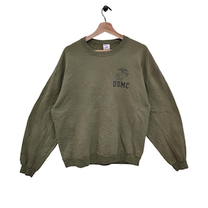 Könnte beinhalten: Olivgrünes Sweatshirt mit langen Ärmeln. Auf der Vorderseite befindet sich eine schwarze Grafik eines Emblems und die Buchstaben "USMC". Das Sweatshirt hängt an einem Holzbügel.