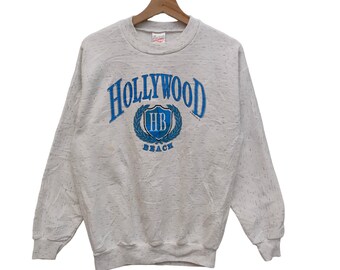 Puede incluir: Una sudadera blanca con un estampado gráfico azul y blanco que dice "HOLLYWOOD" con un escudo que dice "HB" y "BEACH" debajo.