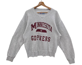Puede incluir: Una sudadera gris con una impresión granate que dice "Minnesota Gophers" con un logotipo granate y blanco en forma de píldora.