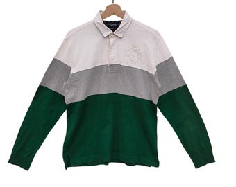 May include: A long-sleeved rugby shirt in white, gray, and green color block design. The shirt has a collared neckline with a three-button placket and an embroidered emblem on the chest. The sleeves are green, and the body is divided into three horizontal color blocks.