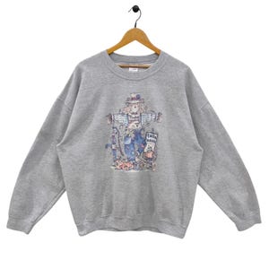 May include: A heather gray sweatshirt featuring a colorful graphic of a scarecrow with a hat, holding a sign that says "fall back". The long-sleeved top is hanging on a wooden hanger.