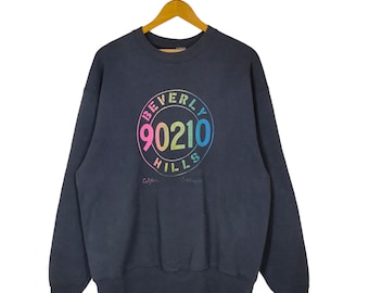 Puede incluir: Sudadera negra con un gráfico de arcoíris de un círculo con el texto "Beverly Hills 90210" en su interior. Los textos "California" y "Los Angeles" están escritos debajo del círculo.