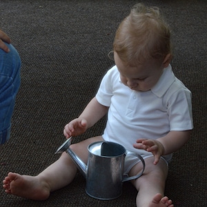 Puede incluir: Un niño pequeño está sentado en una alfombra marrón sosteniendo una regadera plateada. El niño lleva una camisa blanca y tiene el pelo rubio.
