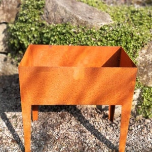 Könnte beinhalten: Ein rechteckiger, rostiger Metall-Pflanzkasten mit vier Beinen. Der Pflanzkasten steht auf einem Kiesbett.