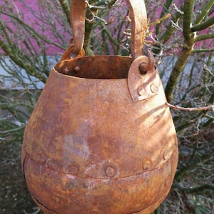 Könnte beinhalten: Ein rostiger Metalleimer mit einem Henkel. Der Eimer ist rund und hat eine strukturierte Oberfläche. Er hat ein abgenutztes, altes Aussehen.