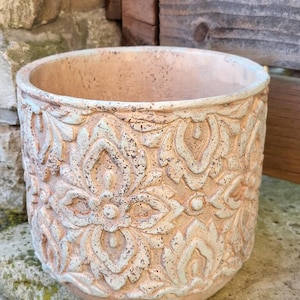 Könnte beinhalten: Runder Blumentopf aus Terrakotta mit detailliertem floralem Muster. Das erhabene Design ist in einem verblassten, hellblauen Farbton gehalten, der einen Kontrast zur Grundfarbe bildet. Geeignet für den Innen- und Außenbereich, ca. 25 cm Durchmesser.