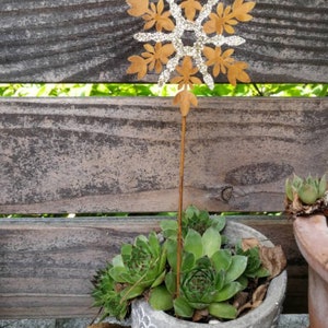 Könnte beinhalten: Ein Metall-Schneeflocken-Ornament mit Glitzerakzenten, platziert in einem Topf mit grünen Sukkulenten. Die Schneeflocke ist an einem langen Metallstiel befestigt.