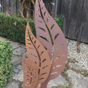 Op de afbeelding: Een groep van drie metalen bladeren sculpturen in verschillende maten. De bladeren hebben uitgesneden aderpatronen en een roestbruine afwerking. De sculpturen zijn gemonteerd op een cirkelvormige basis, ideaal voor tuindecoratie.