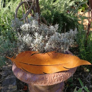 Può includere: Una scultura di piuma in metallo arrugginito con una patina marrone. La scultura è posizionata su un vaso di terracotta con un design decorativo. Il vaso è circondato da piante verdi.