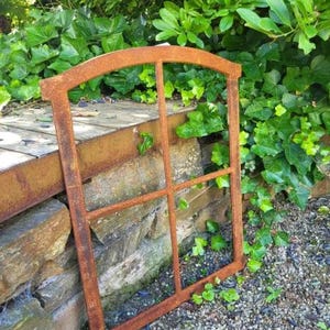 Op de afbeelding: Een verweerde, roestige metalen raamkozijn met een gebogen bovenkant en meerdere ruiten. Het frame staat tegen een achtergrond van weelderig groen gebladerte en een stenen muur, wat een rustieke buitenesthetiek creëert.