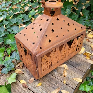 Könnte beinhalten: Eine rechteckige, rostbraune Metalllaterne mit Griff. Die Oberseite hat sternförmige Aussparungen, und die Seiten zeigen eine Silhouette einer Stadt. Die Laterne steht auf einer Holzoberfläche, umgeben von Grün und Herbstlaub.