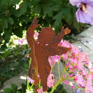 Könnte beinhalten: Ein rostiger Metall-Gartenpfahl in Form eines Cherubins mit ausgestreckten Armen. Der Pfahl ist von rosa Blüten umgeben.
