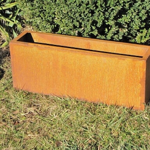 Può includere: Un vaso rettangolare in metallo arrugginito con una finitura marrone invecchiata. Il vaso è vuoto e si trova su un letto di erba verde.