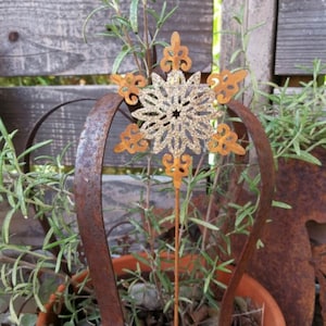 Könnte beinhalten: Rostiges Metall-Gartenornament mit Schneeflocken-Design. Das Ornament befindet sich in einem Terrakotta-Topf mit Grünpflanzen. Im Hintergrund ist ein verwitterter Holzzaun zu sehen, der einen rustikalen Look erzeugt.