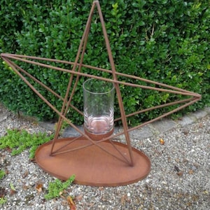 Könnte beinhalten: Ein rostiger, sternförmiger Pflanzer aus Metall mit einem Glaszylinder in der Mitte. Der Pflanzer steht auf einer Kiesfläche.