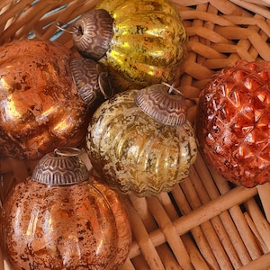 Könnte beinhalten: Eine Sammlung von Vintage-Weihnachtskugeln in einem Weidenkorb. Die Ornamente sind in Gold-, Kupfer- und Orangetönen gehalten und weisen strukturierte Oberflächen und verzierte Metallkappen auf. Der Korb bietet einen rustikalen Hintergrund.