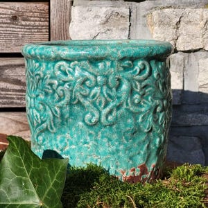 Può includere: Un vaso di fiori in ceramica turchese con finitura craquelé. Il vaso presenta un motivo floreale in rilievo e un aspetto rustico e invecchiato. Il vaso cilindrico poggia su muschio verde.