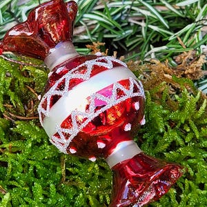 Peut inclure: Ornement de Noël en forme de bonbon, rouge et argenté. L'ornement brillant présente des détails en paillettes blanches et une bande argentée. Il repose sur de la mousse et du feuillage verts.
