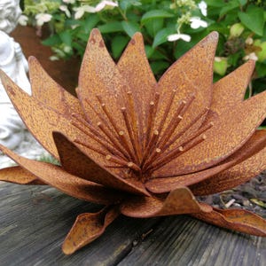 Può includere: Una grande scultura floreale in metallo arrugginito con petali appuntiti e un centro dettagliato. L'opera d'arte da giardino è realizzata in metallo invecchiato, creando un aspetto naturale e invecchiato. Il fiore è posto su una superficie di legno.