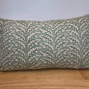 May include: Rectangular decorative pillow with a repeating leaf pattern in shades of green and off-white. The pillow is placed on a wooden surface. The pillow's design features a symmetrical arrangement of stylized leaves.