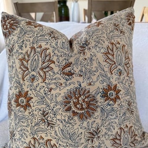 May include: Cream-colored throw pillow with a repeating floral pattern in brown and navy blue. The pillow features large, stylized flowers and intricate leaf designs, creating a vintage or bohemian aesthetic. The pillow is likely made of a soft fabric.