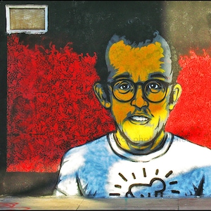 May include: A colorful street art mural of a person wearing glasses and a white t-shirt with a black and white graphic design. The background is red and black.