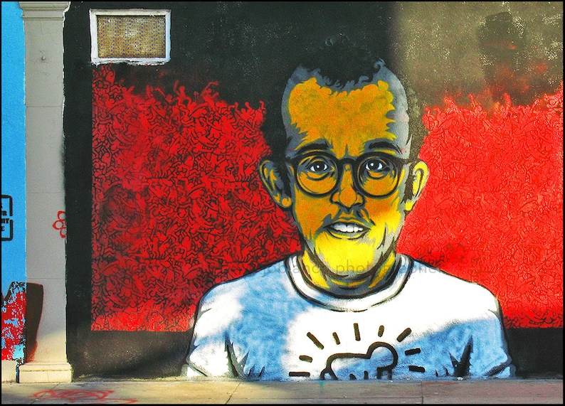 May include: A colorful street art mural of a person wearing glasses and a white t-shirt with a black and white graphic design. The background is red and black.
