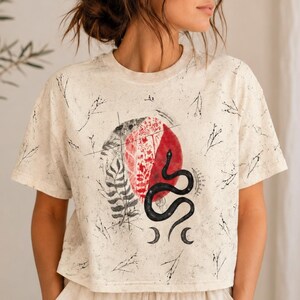 May include: A cream-colored short-sleeve t-shirt with a speckled texture and a black botanical print. The center graphic features a red circular design, a black snake, and moon phases, surrounded by scattered black line drawings of small twigs.