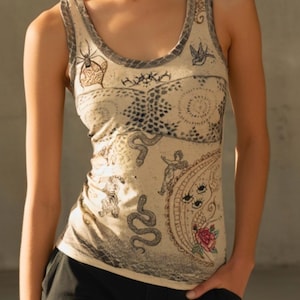 Puede incluir: Una camiseta de tirantes beige con diversas ilustraciones en estilo de tinta negra que incluyen serpientes, una araña, un pájaro, un tigre, un querubín y una luna creciente con ojos. Una rosa roja aparece en la parte inferior derecha de la prenda.
