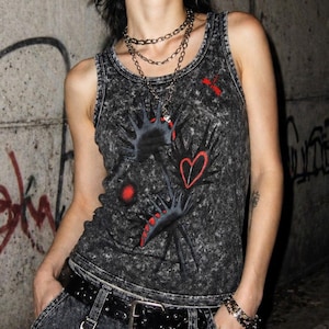 May include: A dark grey tank top with a distressed wash effect. The graphic design features black hand-like shapes with red accents, a red heart, and a red dragonfly. A silver chain necklace is worn over the top.