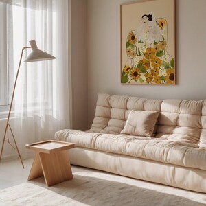 May include: A light beige sofa with a white throw pillow in a living room with a large framed print of a woman with sunflowers. The print has a beige background and the woman is wearing a white dress with sunflowers.