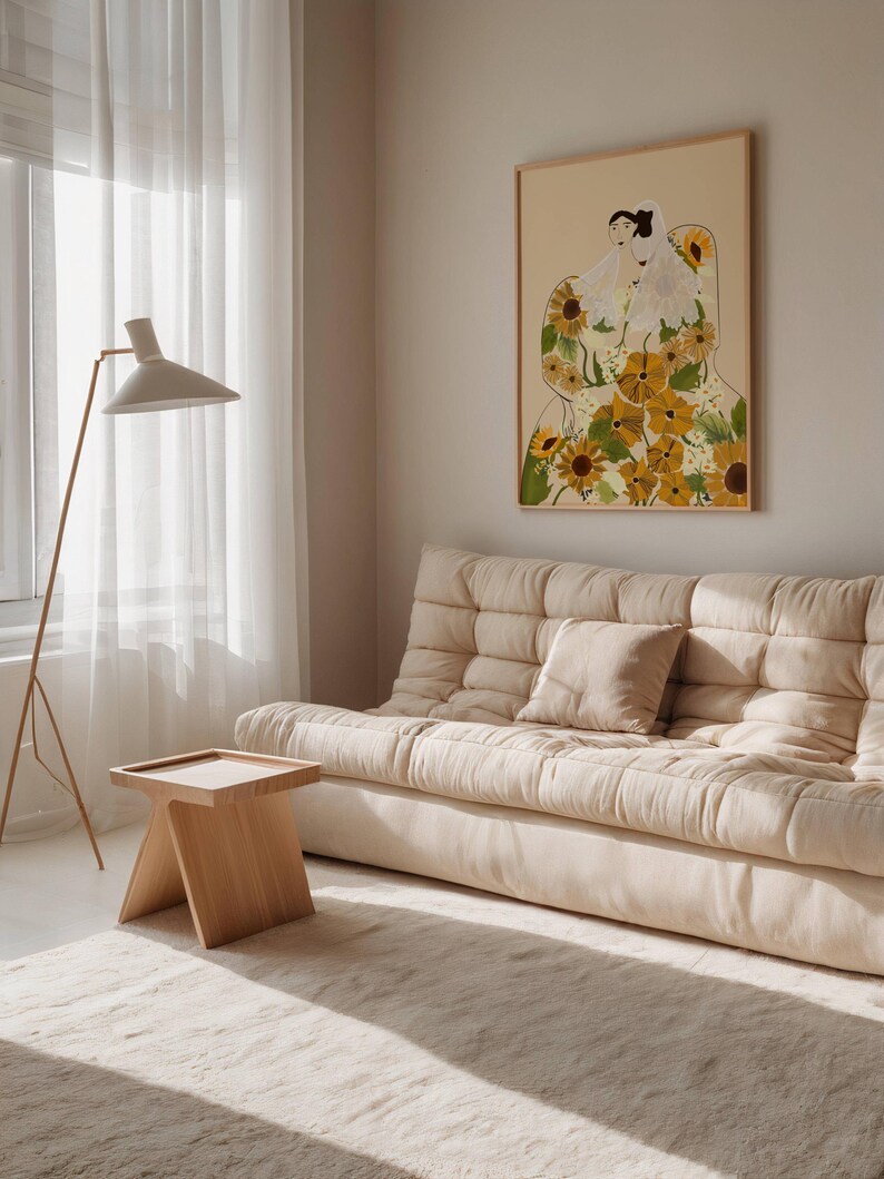 May include: A light beige sofa with a white throw pillow in a living room with a large framed print of a woman with sunflowers. The print has a beige background and the woman is wearing a white dress with sunflowers.