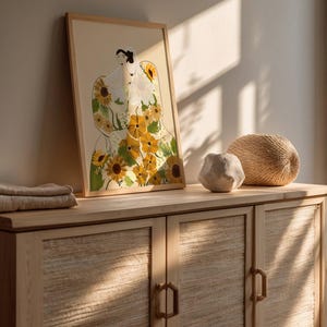 May include: A framed illustration of a person wearing a dress made of sunflowers. The dress is yellow and green with brown stems. The person is wearing a white veil and has dark hair. The frame is light brown wood.