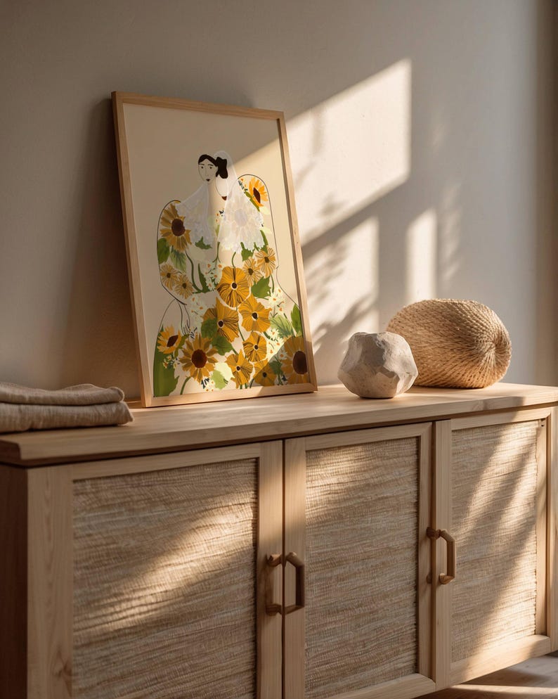 May include: A framed illustration of a person wearing a dress made of sunflowers. The dress is yellow and green with brown stems. The person is wearing a white veil and has dark hair. The frame is light brown wood.